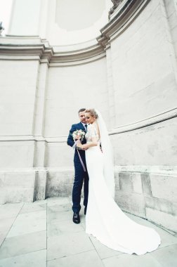 Beyaz gelinlikli sarı saçlı gelin ve beyaz antik bir binanın arka planında takım elbiseli damat. Gelin düğün buketi tutuyor.