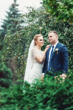 Gelin beyaz gelinlik içinde, bir buket ve parkta poz veren damat..