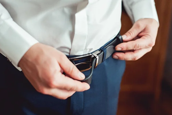 Pengantin pria dengan celana biru dan kemeja putih mengencangkan sabuk kulitnya. calon pengantin pria berkabung. Rincian pernikahan. — Stok Foto