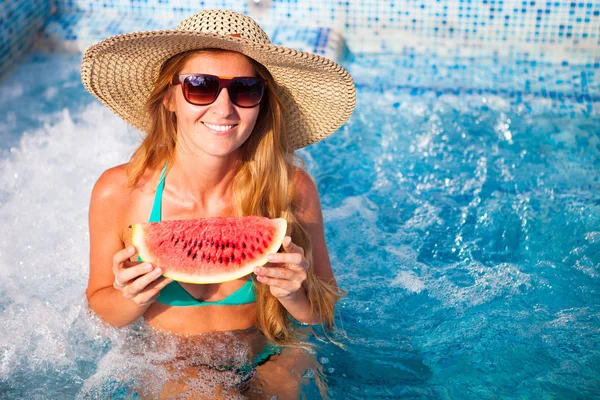 A girl holds half a red watermelon  over a blue pool, relaxing o