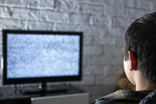 Menino assistindo TV lcd em um quarto escuro com paredes de tijolo estilo loft, vista traseira . — Fotografia de Stock