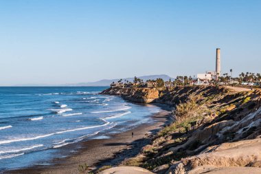 Man walks near cliffs on South Carlsbad State Beach in San Diego, California with the power plant landmark tower in the background.  clipart
