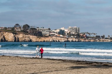 La Jolla, Kaliforniya/ABD - 28 Nisan 2018: Kadın La Jolla Shores Beach, La Jolla Cove, deniz mağaraları ve arka planda bir paddleboarder kıyı boyunca yürümek. 