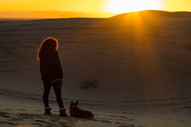 Afrika kökenli Amerikalı bir kadının çöl kumullarında gün batımını izlerken ayağında bir Corgi vardı.. 