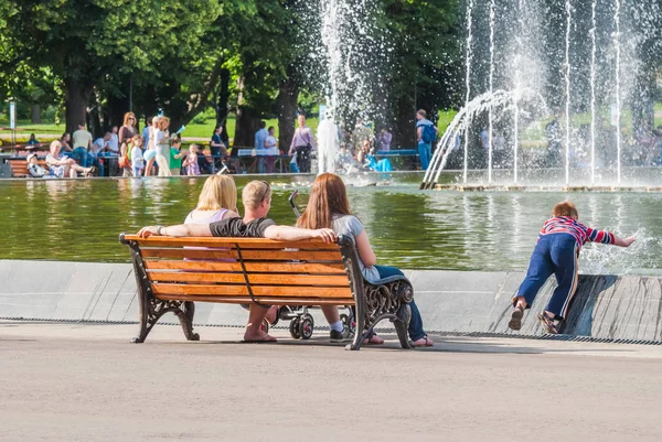 Familia descansando en un día caluroso junto a la fuente en Gorky Park. Un hombre y dos mujeres sentados en el banco, y el niño coge el agua — Foto de Stock