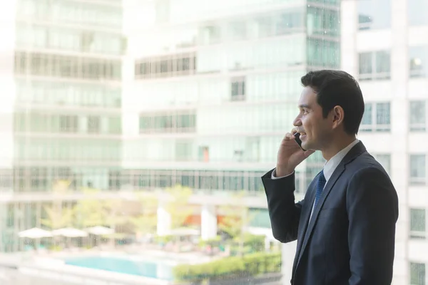 Ejecutivo de negocios maduro y confiado mirando por grandes ventanales a una vista de la ciudad de abajo, desde el último piso de un edificio de oficinas, mientras habla por su teléfono móvil — Foto de Stock