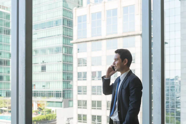 Ejecutivo de negocios maduro y confiado mirando por grandes ventanales a una vista de la ciudad de abajo, desde el último piso de un edificio de oficinas, mientras habla por su teléfono móvil — Foto de Stock