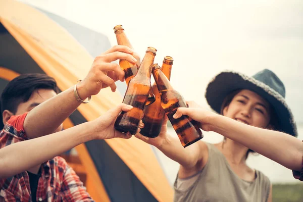 Group of man and woman enjoy camping picnic and barbecue at lake with tents in background. Young mixed race Asian woman and man. Young people's hands toasting and cheering beer. Vintage filtered image