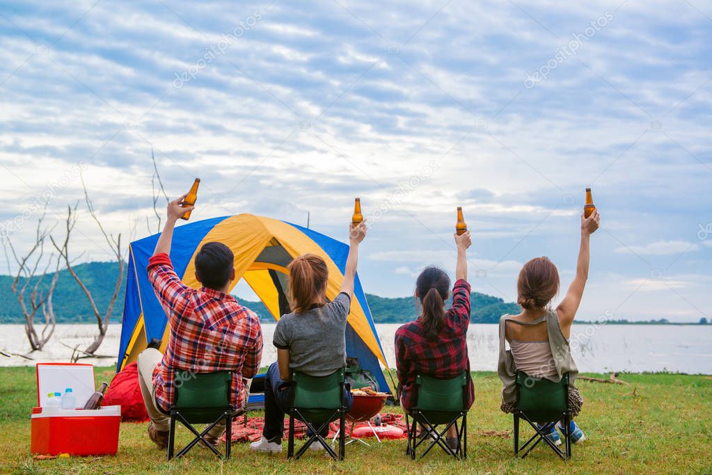 Group of man and woman enjoy camping picnic and barbecue at lake with tents in background. Young mixed race Asian woman and man.