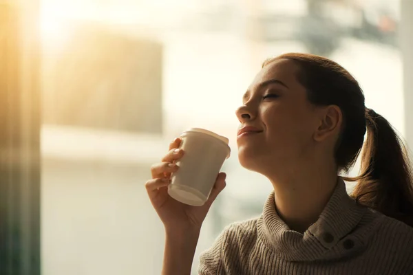 Frau trinkt Kaffee zu Hause, während Sonnenaufgang durch Fenster einströmt und Fackeln in die Linse erzeugt. — Stockfoto