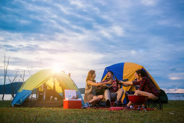 Group of man and woman enjoy camping picnic and barbecue at lake with tents in background. Young mixed race Asian woman and man. Young people's hands toasting and cheering bottles of beer. — Stock Photo, Image