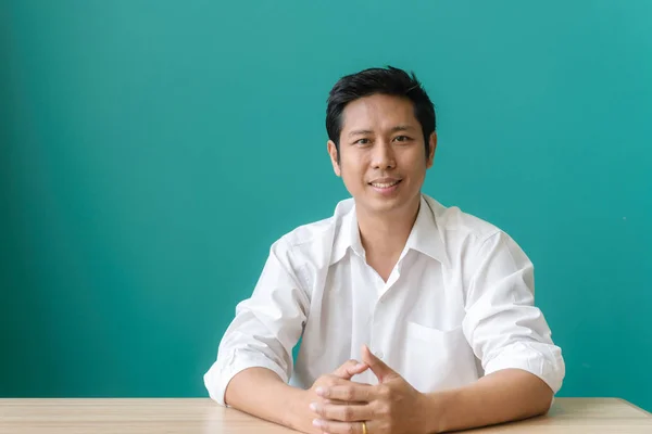 Jobber med glede. Portrett av asiatisk forretningsmann som smiler og ser på kamera med smil mens han sitter på arbeidsplassen sin med blå vegg. Vintage-filtrert bilde . – stockfoto