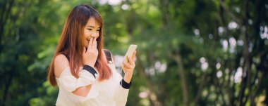 Şirin Asyalı kadın sıcak bahar günü, kulaklık müzik dinlemek çok güzel kadın parkta otururken hoş kısa mesaj cep telefonu sesleri okuyor. Panoramik afiş.