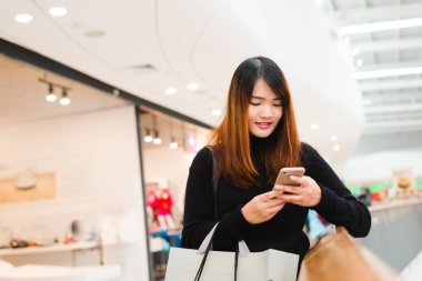 Alışveriş Merkezi, güzel genç Asyalı kadın kapalı ağ için akıllı telefon kullanarak gülümseyen portresi. Teknoloji, seyahat yaşam biçimi kullanan turist kadın. Alışveriş Merkezi dış giyim mağazası ile.