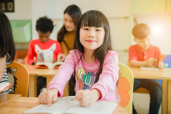Asiatisk jente leser en bok som smiler til kameraet. Rekke med multietniske elementære studenter leser bok i klasserommet på skolen. Bilder av årgangseffekt . – stockfoto