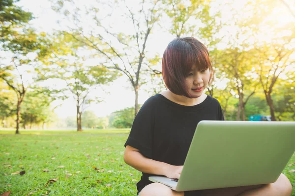 Ung asiatisk kvinnes bein på det grønne gresset med åpen laptop. Jentas hender på tastatur. Fjernunderingskonsept. God hipster-ung asiatisk kvinne som jobber med laptop i parken. Student som studerer utendørs . – stockfoto