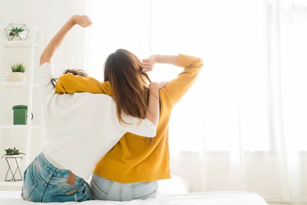 Back view of women lesbian happy couple waking up in morning, sitting on bed, stretching in cozy bedroom, looking through window. Funny women after wakeup. Lesbian couple together indoors concept. — Stock Photo, Image