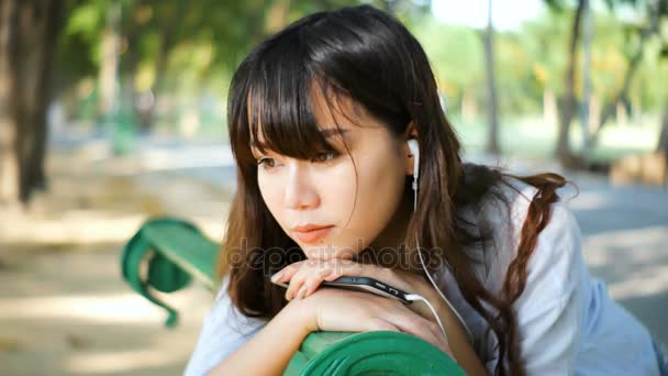 美丽的年轻的时髦的悲伤的亚洲妇女情绪听音乐在耳机与智能手机 坐在公园的长凳在夏天夕阳阳光 — 图库视频影像
