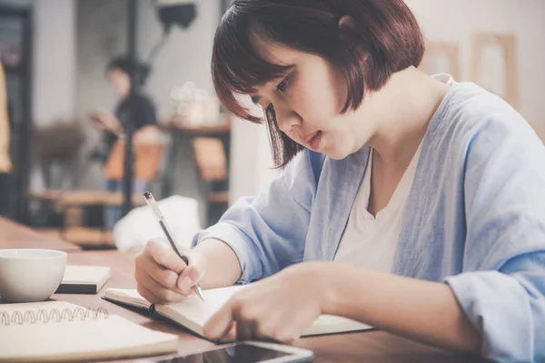 Młoda kobieta w białej sukni, siedząc przy stoliku w kawiarni i pisanie w notesie. Asian kobieta rozmawia smartphone i filiżankę kawy. Freelancer pracuje w kawiarni. Vintage efekt stylu Zdjęcia — Zdjęcie stockowe