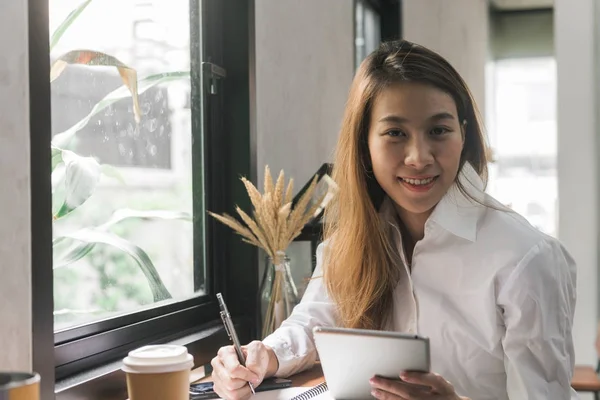 En ung forretningskvinne i hvit kjole sitter ved bordet i kafeen og skriver i en notatbok. Asiatisk kvinne bruker tablett og kopp kaffe. Freelancer jobber på kafeen. Student læring på nett . – stockfoto
