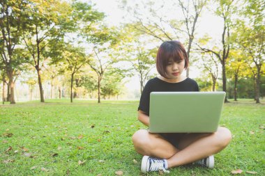 Genç Asyalı kadının bacakları açık dizüstü bilgisayar ile yeşil çimenlerin üzerinde. Kızın eller klavye. Mesafe öğrenme kavramı. Park içinde laptop üzerinde çalışan mutlu hipster genç Asyalı kadın. Öğrenci açık havada.