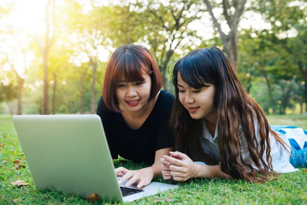 Jovens mulheres asiáticas deitadas na grama e usando laptop e digitação. Meninas mãos no teclado. Conceito de ensino à distância. feliz hipster jovem asiático mulheres trabalhando no laptop no parque. Estudante estudando ao ar livre . — Fotografia de Stock