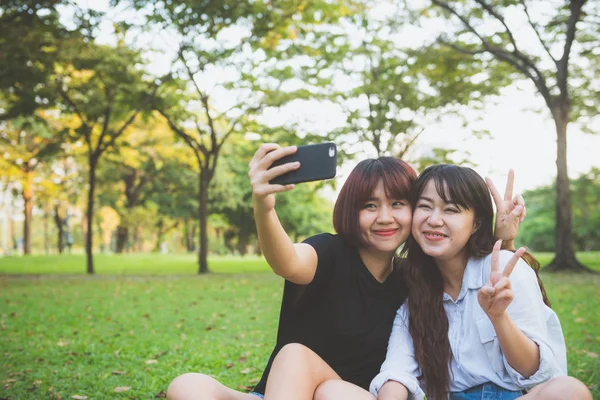 To vakre, lykkelige, unge asiater som har det gøy sammen i parken og tar en selfie. Glade, unge asiater som smiler og ser på smarttelefonen. Livsstil og vennskapskonsepter . – stockfoto