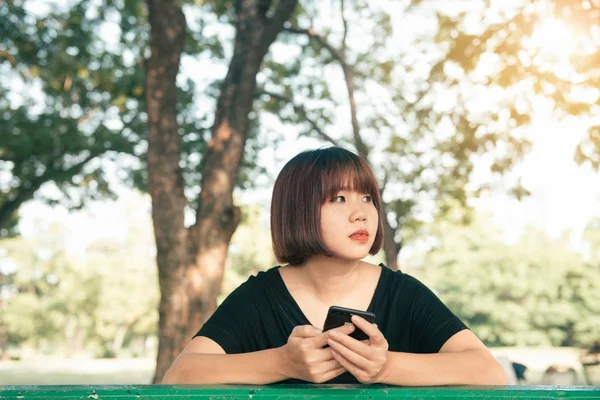 Linda mujer asiática leyendo agradable mensaje de texto en el teléfono móvil mientras está sentado en el parque el día de primavera. Mujer asiática usando en el teléfono inteligente con sensación de relax y la cara sonriente. Conceptos de estilo de vida y tecnología . —  Fotos de Stock