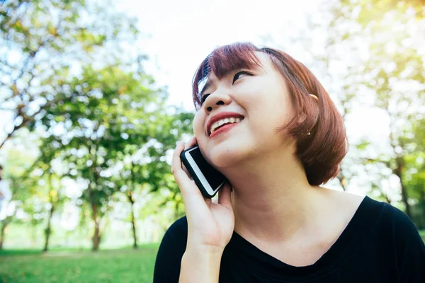 Søt asiatisk kvinne smiler og snakker i mobiltelefon mens hun sitter i parken på vårdagen. Asiatisk kvinne som bruker smarttelefon med følelse av å slappe av og smile. Livsstil og teknologiske konsepter . – stockfoto