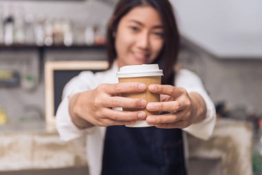 Asyalı kadın barista counter arka plan ile çevrili gülümseme ile müşterisi için bir fincan kahve hizmet tutun bir genç kapatın. Genç kadın barista ve onun küçük bir dükkanda. Yiyecek ve içecek konsepti