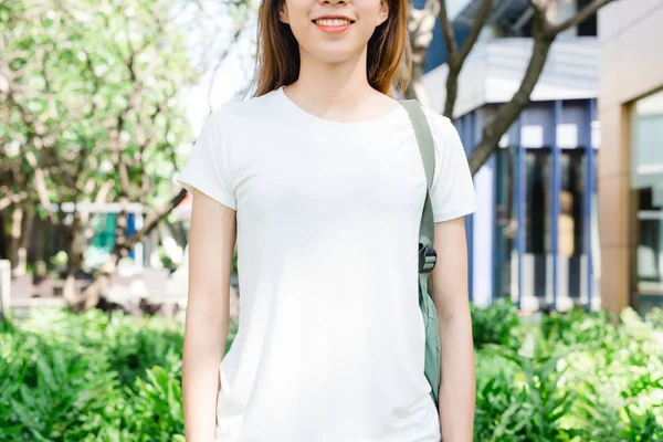 Asiatisk hipsterjente langt brunt hår i hvit, blank T-skjorte står midt i gata. En kvinne i gateklær står på grønn bybakgrunn. Tomme mønstringsområder for tekst eller design . – stockfoto