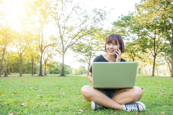 Søt asiatisk kvinne smiler og snakker i mobiltelefon mens hun sitter i parken på vårdagen. Asiatisk kvinne som bruker smarttelefon og laptop med følelse av å slappe av og smile. Livsstil og teknologiske konsepter . – stockfoto