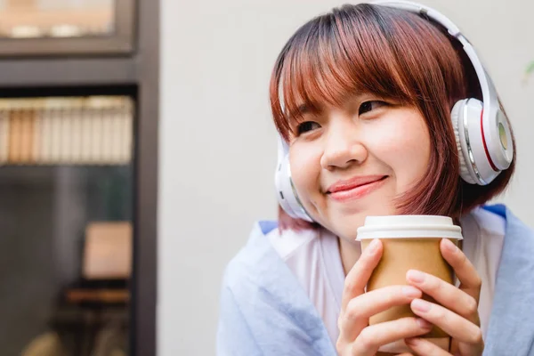 Feliz joven asiática escuchando música en una cafetería con una taza de café en la mano. Mujer joven escuchando música con auriculares mientras se relaja en el jardín. Café relajar concepto . —  Fotos de Stock