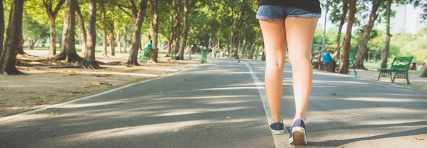 Asian woman's legs while jogging in the park covered with plenty of trees and warm sunlight. Young woman legs close up when running in the park. Outdoor activity in the park concept. Panoramic banner. — Stock Photo, Image