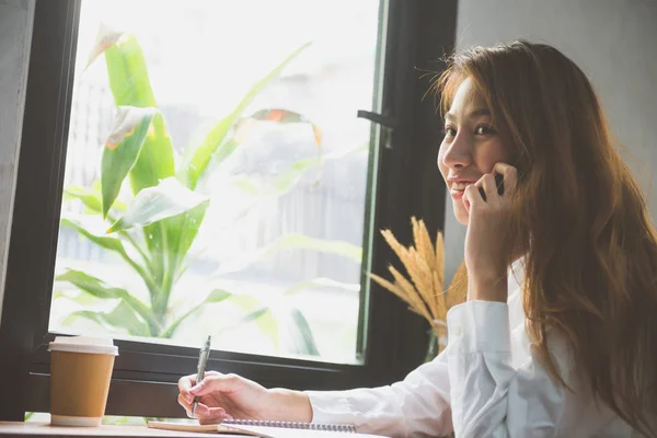 Joven mujer de negocios vestida de blanco sentada a la mesa en la cafetería y escribiendo en un cuaderno. Mujer asiática hablando smartphone y taza de café. Freelancer trabajando en una cafetería. Aprendizaje del estudiante en línea . —  Fotos de Stock