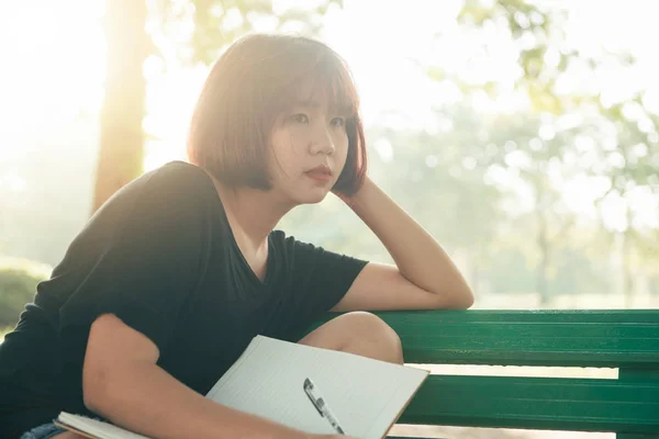 Feliz hipster joven asiática mujer escribiendo en su diario en el parque. Feliz hipster joven asiática mujer trabajando en el cuaderno en el parque. Estudiante estudiando al aire libre. Estilo de vida concepto al aire libre mujer. Luz de bengala . —  Fotos de Stock