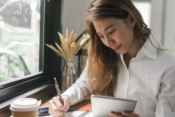 Nuori nainen valkoisessa puvussa istuu pöydässä kahvilassa ja kirjoittaa muistikirjaan. Aasialainen nainen käyttää tabletin ja kupin kahvia. Freelancer työskentelee kahvilassa. Opiskelijan oppiminen verkossa . — kuvapankkivalokuva