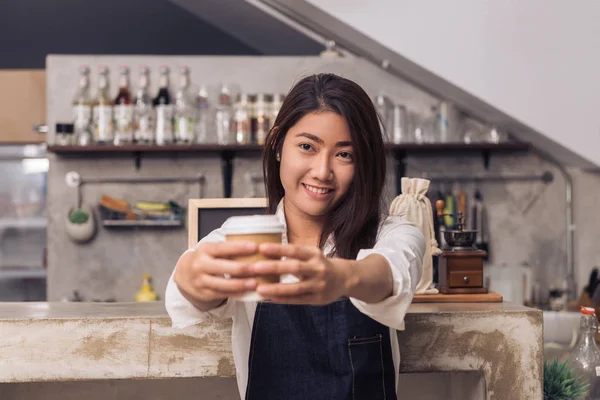 Lähikuva nuori aasialainen nainen barista pitää kupin kahvia palvelevat asiakkaalleen hymyillen ympäröi baari laskuri tausta. Nuori barista ja hänen pieni liikkeensä. Elintarvikkeiden ja juomien käsite — kuvapankkivalokuva