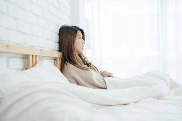Happy beautiful young Asian woman waking up in morning, sitting on bed, stretching in cozy bedroom, looking through window. Funny asian woman after wake up. Asia woman is stretching and smiling. — Stock Photo, Image
