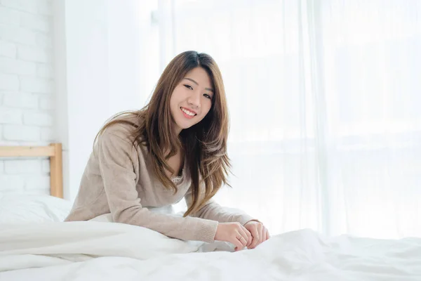 Onnellinen kaunis nuori aasialainen nainen herää aamulla, istuu sängyllä, venyttely viihtyisässä makuuhuoneessa, katselee ikkunasta. Hauska aasialainen nainen heräämisen jälkeen. Aasia nainen on venyttely ja hymyilee . — kuvapankkivalokuva