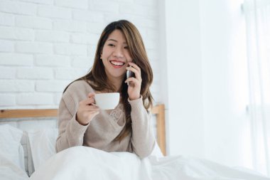 Asyalı kadın sabah kahve tutarken yatağında smartphone kullanarak. Telefon yatak odasında konuşurken ve sıcak kahve zevk güzel Asyalı kadın. yaşam tarzı Asya kadın kavramı