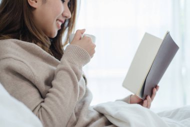 Yatak odasında yatakta kitap okuma ve sıcak kahve zevk güzel Asyalı kadın. Bir kitap ve coffee.lifestyle Asya kadın fincan ev concept tutarak rahat kazak giyen Asya erkek.