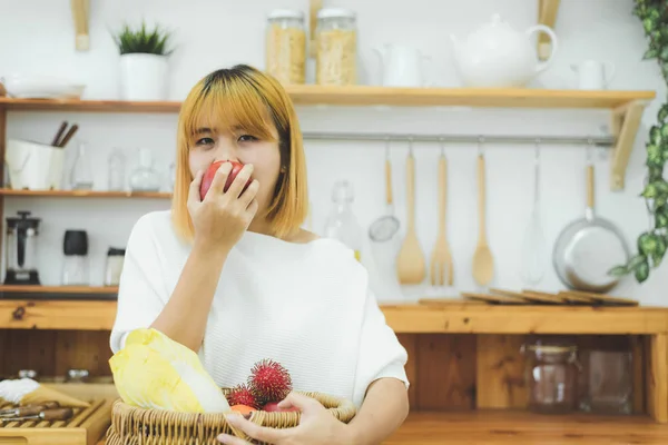 Asiatisk vakker kvinne spiser rødt eple på kjøkkenet hjemme hos seg. Lykkelig, søt asiatisk kvinne spiser frisk frukt for en sunn kropp. livsstil asiatisk kvinne som hjemmebegrep . – stockfoto