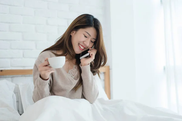 Femme asiatique utilisant le smartphone sur son lit tout en tenant une tasse de café le matin. Belle femme asiatique appréciant le café chaud et parlant au téléphone dans sa chambre. style de vie asiatique femme concept — Photo