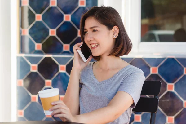 Onnellinen hymy aasialainen liike naiset käyttävät puhuva matkapuhelin istuu kahvilassa ja pitämällä kuppi kahvia. Kaunis iloinen aasialainen nainen kahvilassa puhuu puhelimessa ja hymyilee nauttien kahvista . — kuvapankkivalokuva
