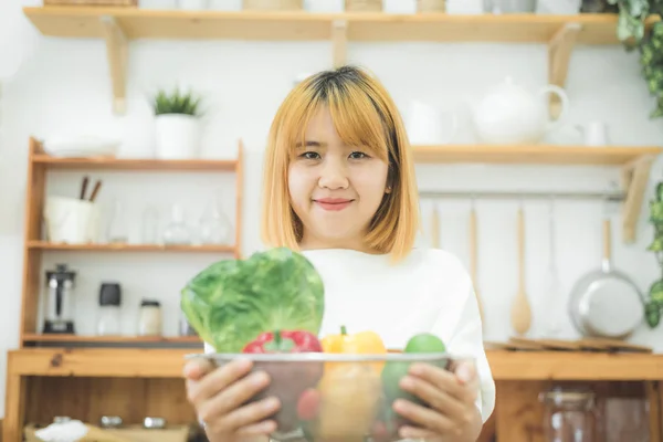 Hymyilevä aasialainen kaunis nainen istuu keittiössä kotona ja pitää salaattikulhoa. Nuori onnellinen nainen syö terveellistä salaattia vihreillä tuoreilla ainesosilla terveelle keholle . — kuvapankkivalokuva