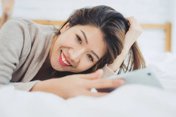 Lykkelige asiatiske kvinner bruker smarttelefon i senga om morgenen. Asiatisk kvinne i sengen sjekker sosiale apper med smarttelefon. Smilende surfenett med mobiltelefon hjemme. Mobilavhengig konsept . – stockfoto