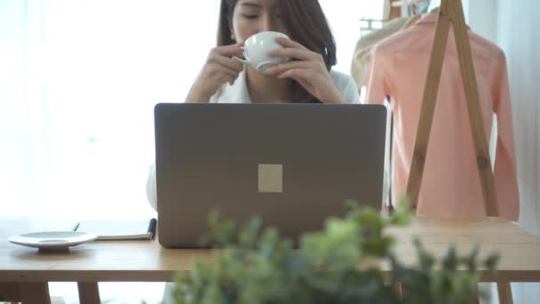 Slow motion - krásná mladá usmívající se žena pracující na notebooku zároveň pití teplé kávy sedí v obývacím pokoji doma. Se těší času doma. Asijské ženy pracující ve své domácí kanceláři