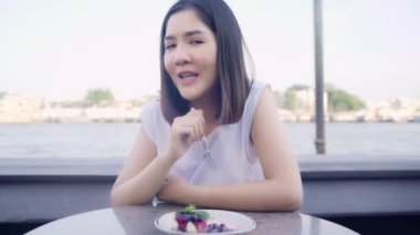 Blogger Asyalı arkadaş canlısı kadın etkileyicisi kafede pasta yiyor. Genç bayan, mutlu bir şekilde rahatlayın. Şehirdeki nehrin yakınındaki sosyal medya tatil seyahatinde video kaydını kullanarak rahatlayın..