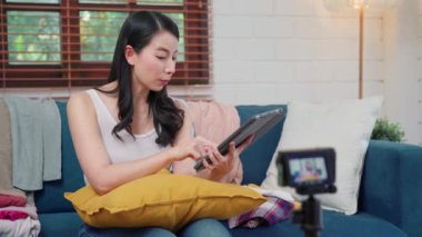 Asyalı moda tasarımcısı kadınlar kamera yayınını kullanıyor ve giysi ticareti satmak için canlı yayın yapıyor, tablet sohbet kullanarak e-ticaret yayını yapıyor evdeki oturma odasındaki koltukta oturan müşteriye cevap veriyor..